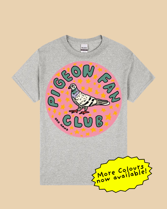 A heather gray t-shirt featuring a circular graphic with a pigeon and the text 'PIGEON FAN CLUB' in green and pink, displayed on a beige background with a yellow 'More Colours now available!' sticker overlay.