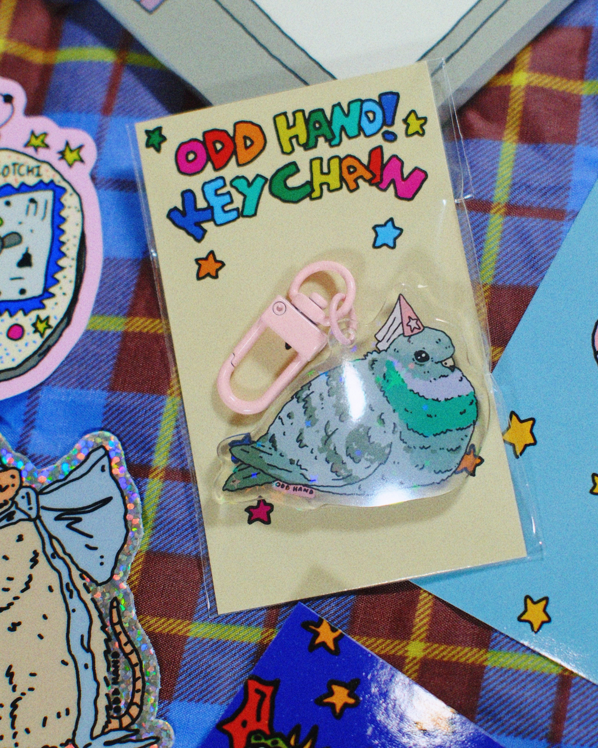 A packaged keychain featuring a cartoon pigeon character with the text 'ODD HAND! KEYCHAIN' on a cream backing card, placed on a plaid fabric surface with other colorful items partially visible around it.