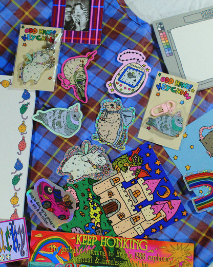 A collection of colorful psychedelic and cartoon-themed stickers and patches arranged on a blue and red plaid fabric background. The designs include a rabbit, a castle, a toucan, a camo-patterned cat, and other whimsical illustrations with text such as 'God Happy Cuddly' and 'Keep Honking'.